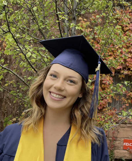 Headshot of white female with blonde and brown hair smiling in a navy blue and yellow cap and gown in front of a tree with green and orange leaves.